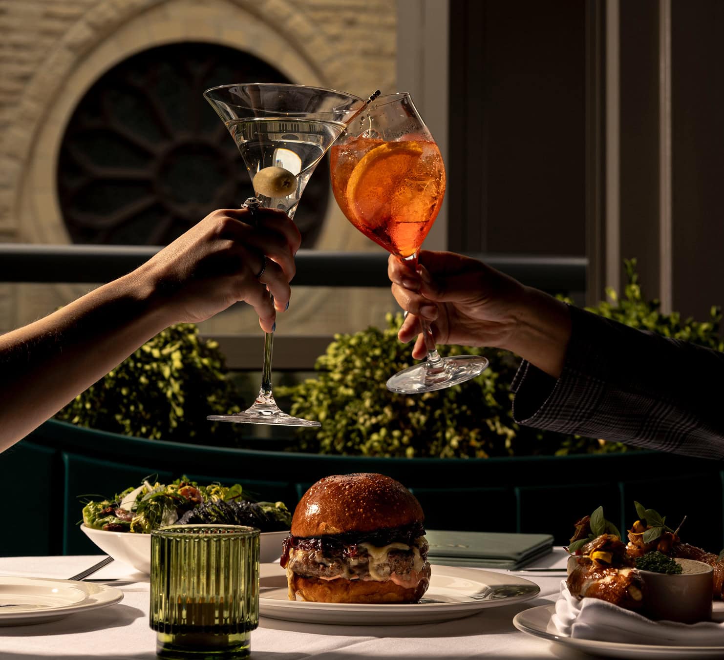 Two guests of the alston tapping glasses overtop of a cheese burger and other food on a table.