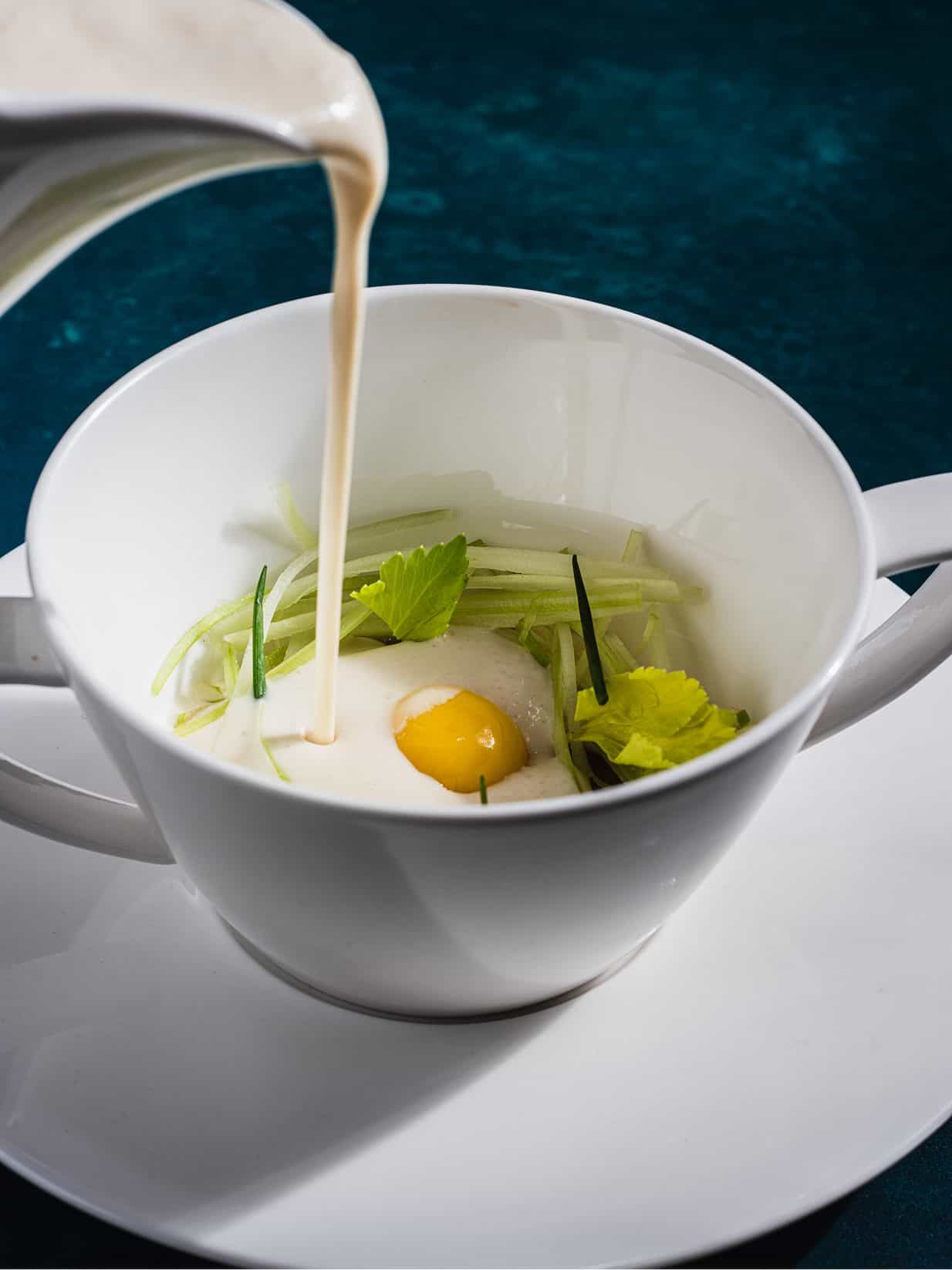Eggs Benedict with fresh greens and creamy sauce in a white cup.